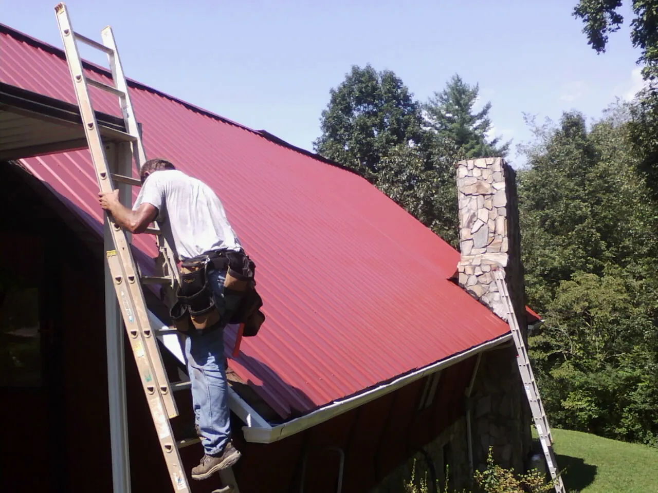 Roofer on a ladder beside a red metal roof and stone chimney — Soffit & Fascia Repair by Roofing Brothers in Rogers